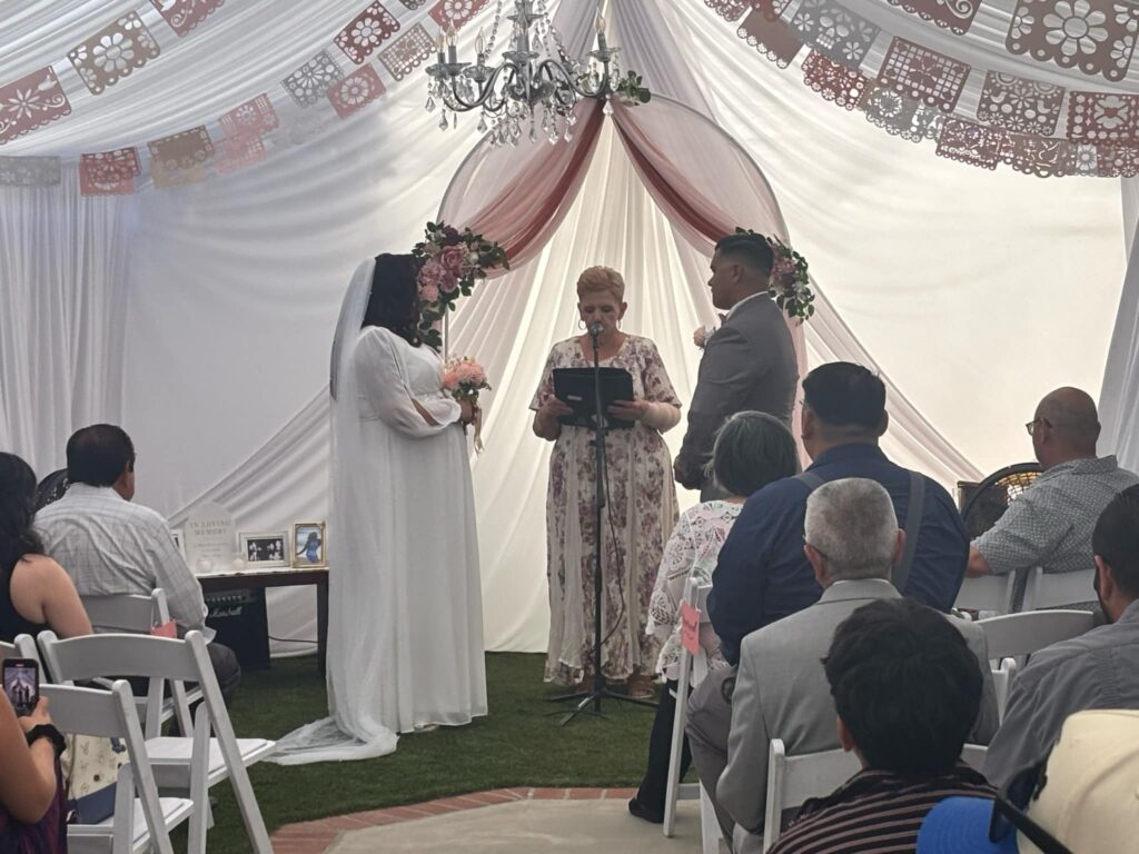 Wedding ceremony scene with a bride and groom standing before an officiant, surrounded by guests in a decorated venue, featuring floral arrangements and elegant drapery.